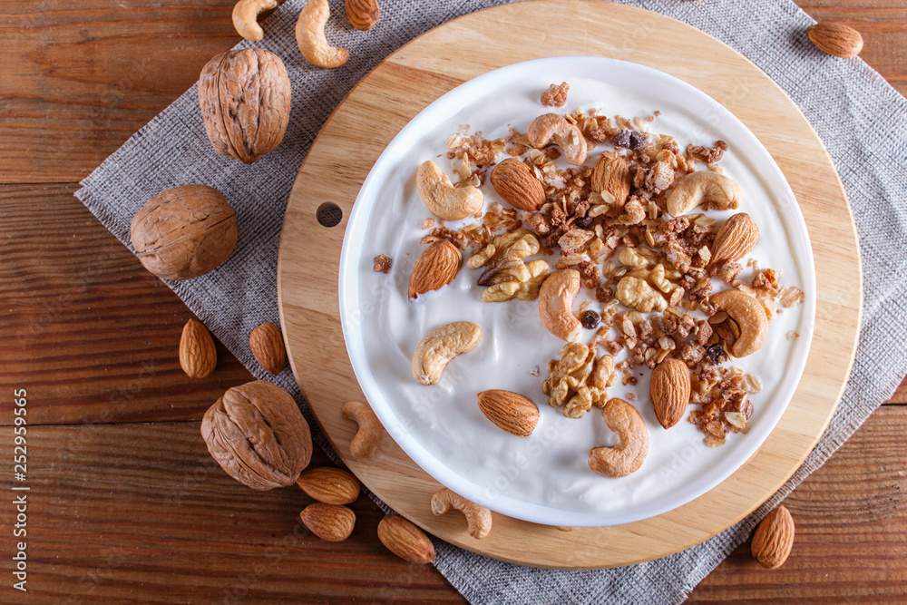 White plate with greek yogurt, granola, almond, cashew, walnuts  on brown wooden background.