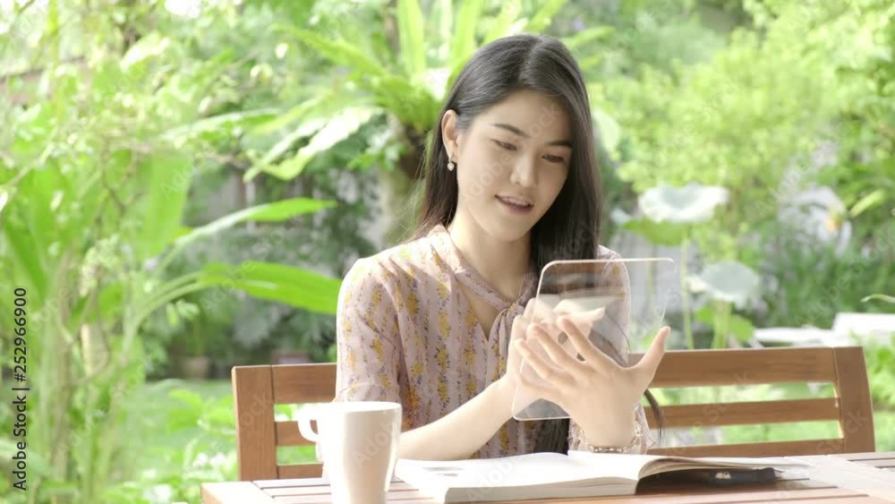 Young beautiful Asian woman browsing news feed with high tech glass display in her home garden with green tree in background and drinking coffee. Future technology concept. For graphic overlay. Young beautiful Asian woman browsing news feed with high tech glass display in her home garden with green tree in background and drinking coffee. Future technology concept. For graphic overlay.