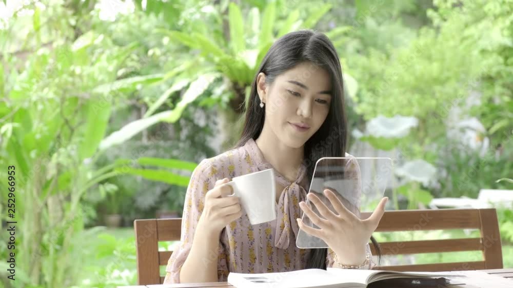 Young beautiful Asian woman browsing news feed with high tech glass display in her home garden with green tree in background and drinking coffee. Future technology concept. For graphic overlay.