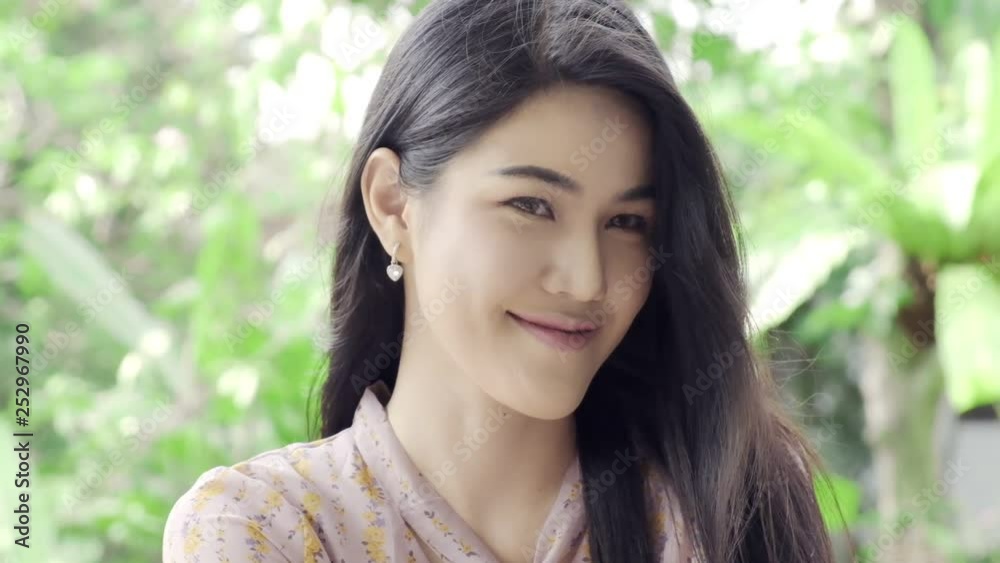 Super Slow motion of young beautiful Thai woman smiling at camera in her home garden with green tree in background, close up shot. Millennial lifestyle concept. Super Slow motion of young beautiful Thai woman smiling at camera in her home garden with green tree in background, close up shot. Millennial lifestyle concept.