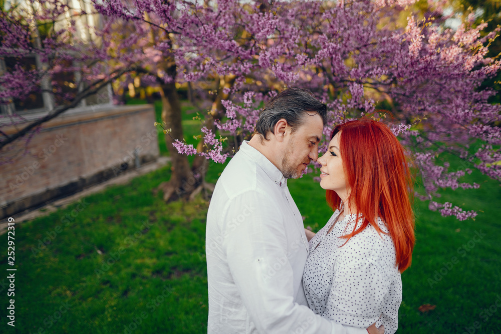 beautiful and stylish redhead woman in a white blouse standing in a sunny summer park with her handsome husband near flower trees