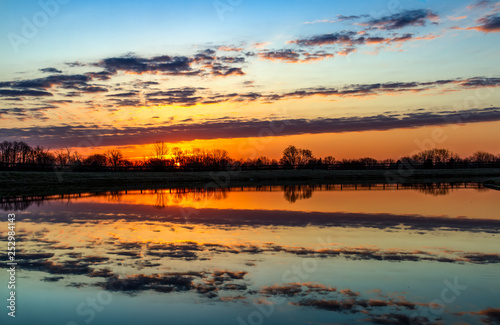 Country Pond Morning - The colors of the rising sun and clouds are reflected on a country pond with a fence surrounding it.