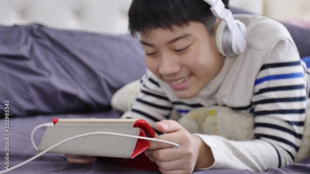 UHD Dolly shoot of asian cute boy watching on tablet computer and wearing white headphone with smile face. UHD Dolly shoot of asian cute boy watching on tablet computer and wearing white headphone with smile face.