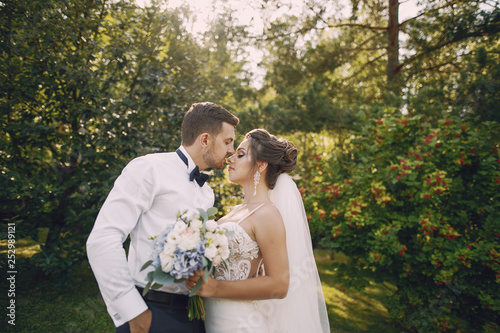 A young and beautiful bride and her husband is standing in a summer park with bouquet of flowers photo