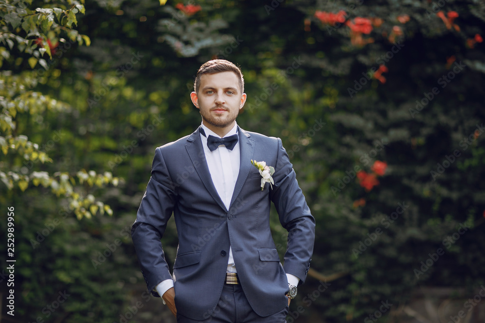 A handsome and stylish bride in a blue suit stands in a summer park near the bushes with bouquet A handsome and stylish bride in a blue suit stands in a summer park near the bushes with bouquet