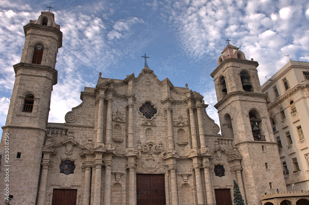 Facade of cathedral in Havana, Cuba with sandstone towers and brass bells and cross symbols on roof under bright blue sky and white clouds