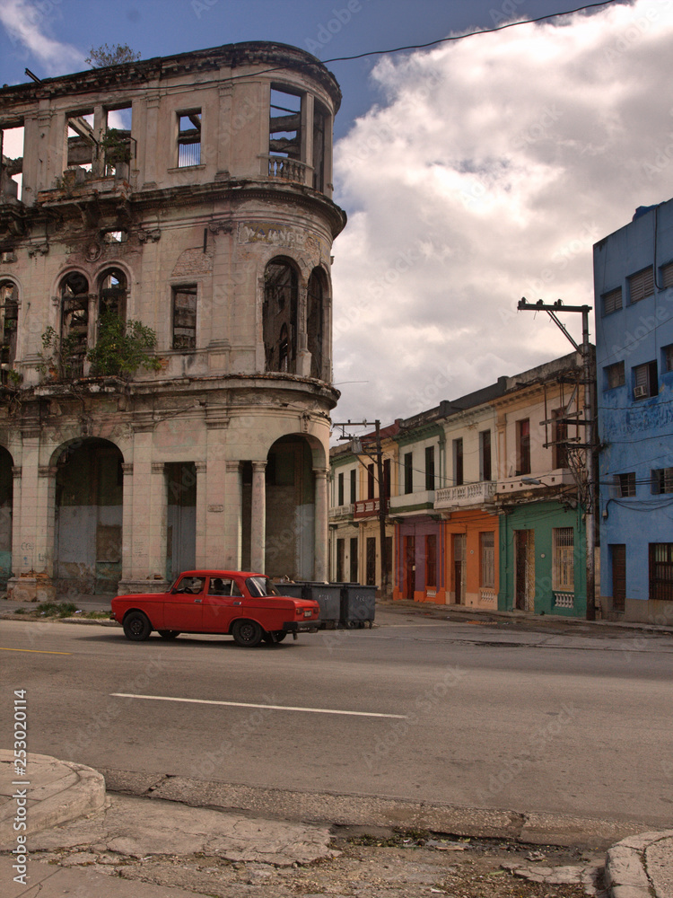 Facade of classic old heritage building under bright blue sky and white clouds in downtown Havana, Cuba with plants growing from window, red car and road in foreground, and colorful row houses in back