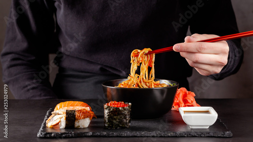 Concept of Asian cuisine. The girl is holding Japanese chopsticks in her hand and eating Chinese noodles from a black plate in a restaurant. background image. copy space