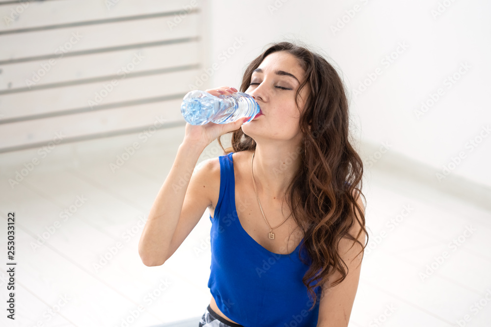 Healthy lifestyle, people and sport concept - Smiling woman sitting on yoga mat and drinking water after hard workout Healthy lifestyle, people and sport concept - Smiling woman sitting on yoga mat and drinking water after hard workout