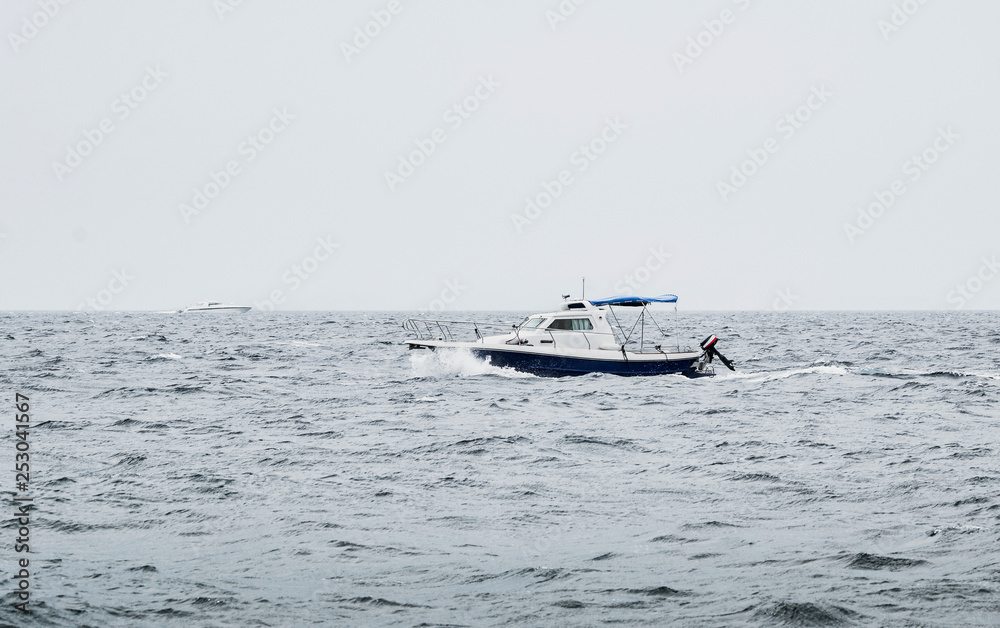 A motorboat floating on the heavy sea with waves in bad weather conditions with other luxury yacht on background. Boat on the ocean (overcast day) after the storm - image A motorboat floating on the heavy sea with waves in bad weather conditions with other luxury yacht on background. Boat on the ocean (overcast day) after the storm - image