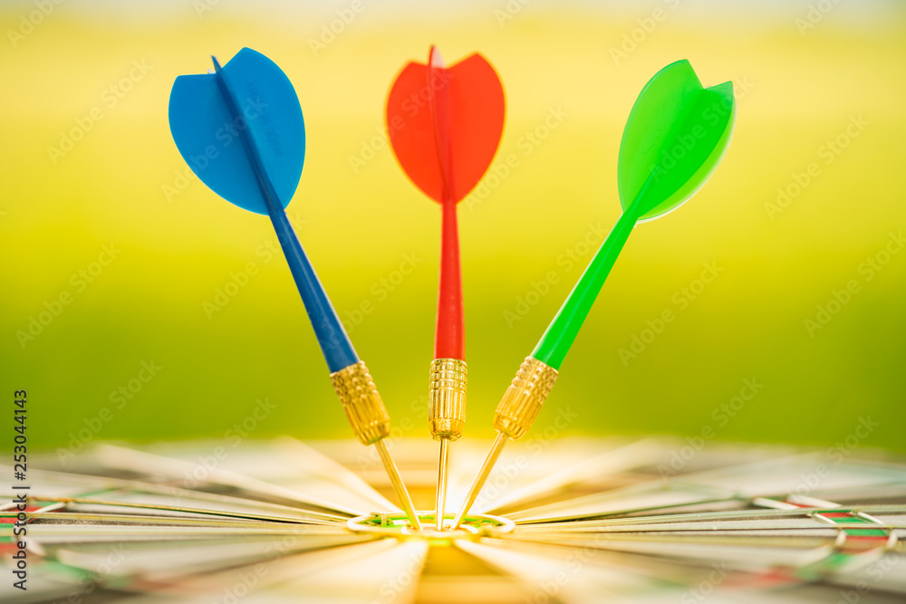 Red, Green and Blue dart arrows hitting in the target center of dartboard with green nature background. Target business, achieve and victory concept. Red, Green and Blue dart arrows hitting in the target center of dartboard with green nature background. Target business, achieve and victory concept.