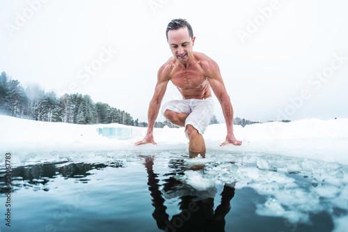 Young man with lean muscular body going to swim in the cold winter water with ice floating on the surface and forest on the background