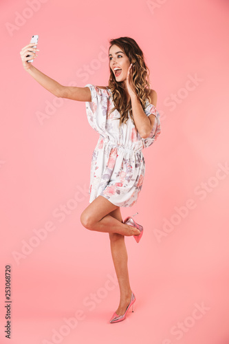 Full length image of european woman 20s with long hair smiling while taking selfie on mobile phone