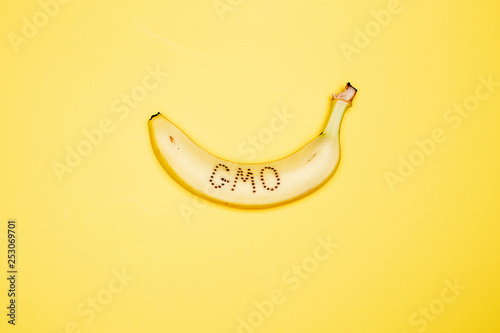 Ripe banana on the peel of which is written the word "GMO" lies on a bright yellow background.