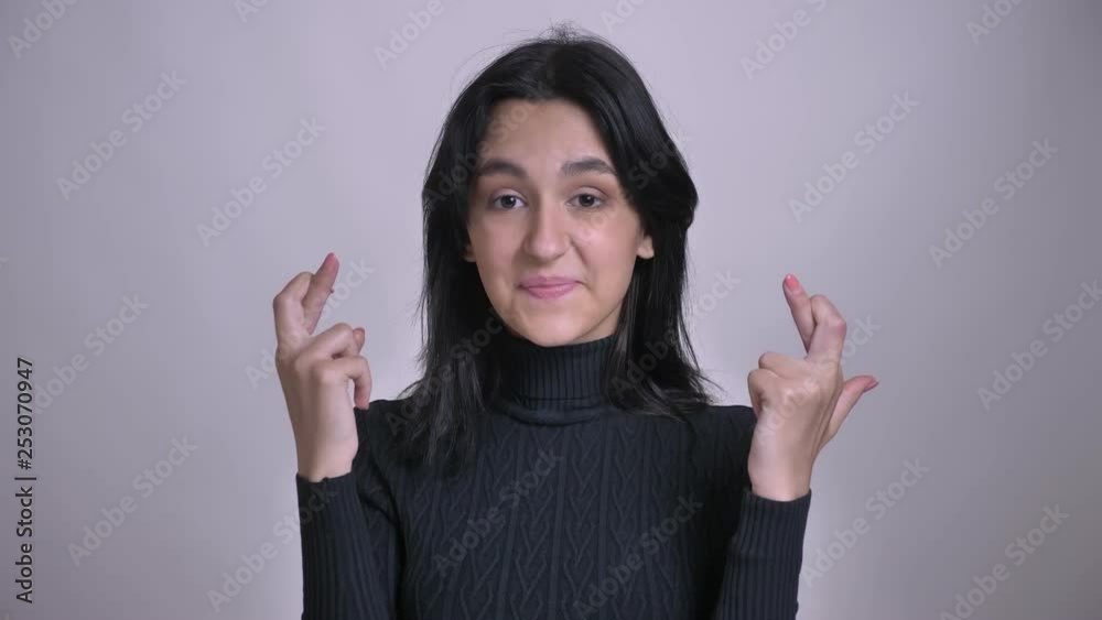 Closeup shoot of young pretty caucasian female having her fingers crossed in hope while looking anxiously at camera