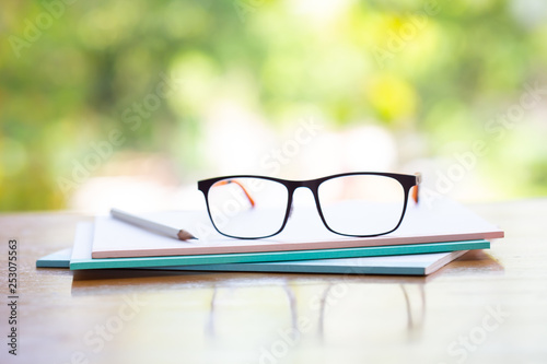 Black eyeglasses, white pencil with three white notebook on wooden table, Bokeh garden background, Close up & Macro shot, Selective focus, Stationery concept