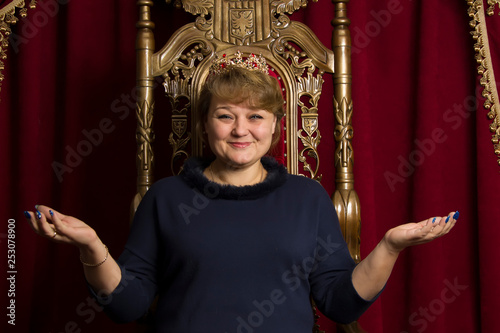A middle-aged woman wearing a crown in a beautiful armchair. Ordinary woman queen.woman in a red chair