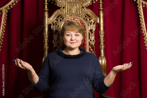 A middle-aged woman wearing a crown in a beautiful armchair. Ordinary woman queen.woman in a red chair