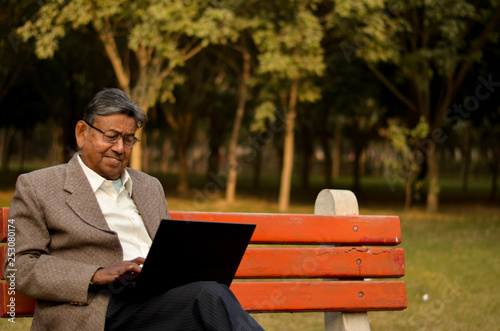 Close up of a senior Indian man wearing a tweed coat working on a laptop on a park bench in Delhi, India