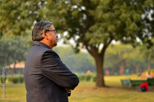 Side pose of a senior Indian man wearing a suit and looking away into distance standing and smiling in a park in Delhi, India