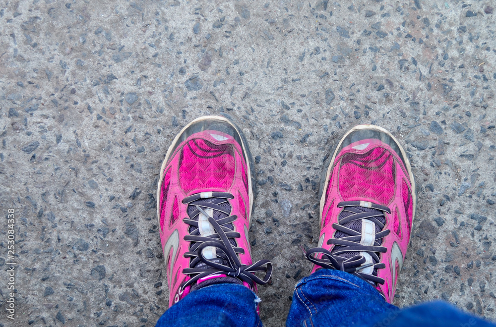 This is a top view of pink colored sports shoes with tied laces. the shoes are worn by a girl woman standing on a tarmac road also wearing blue jeans or trousers This is a top view of pink colored sports shoes with tied laces. the shoes are worn by a girl woman standing on a tarmac road also wearing blue jeans or trousers