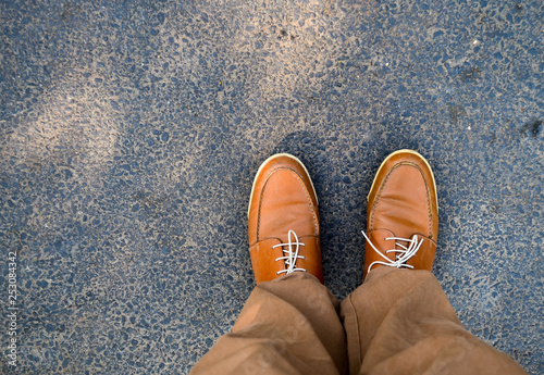 This is a top view of brown camel colored Brogue leather shoes with tied laces. the shoes are worn by a person standing on a tarmac road also wearing brown/khaki colored pants or trousers