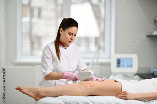Beautiful doctor cosmetician performs a cosmetic procedure on the patient hips with electronic medical equipment in beauty parlor