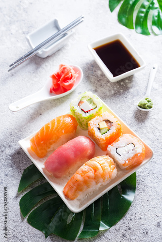 Various sushi on ceramic plate with metal Korean sticks on light stone background with green leaves, flat lay