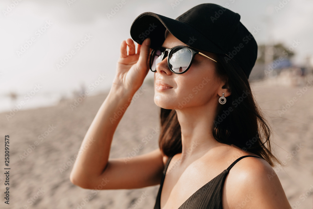 Stylish modern woman in trend outfit looking on the ocean with happy smile wearing cap and glasses and enjoys summer hot days. Young caucasian female model on the sea shore Stylish modern woman in trend outfit looking on the ocean with happy smile wearing cap and glasses and enjoys summer hot days. Young caucasian female model on the sea shore