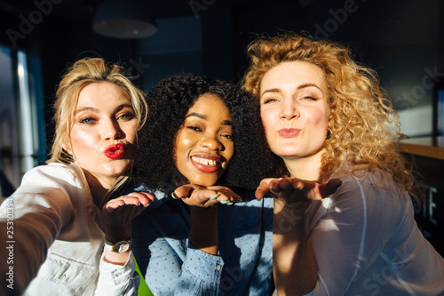 Self portrait of funky childish trio gesturing kiss by duck face shooting selfie on front camera having fun, hugging on dark background. Rest relax leisure concept