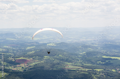 Lone paraglider gliding through the air over a green, mountainous backdrop and partially clouded skies.