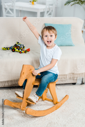Photography adorable excited preschooler boy riding rocking horse and gesturing with hand at