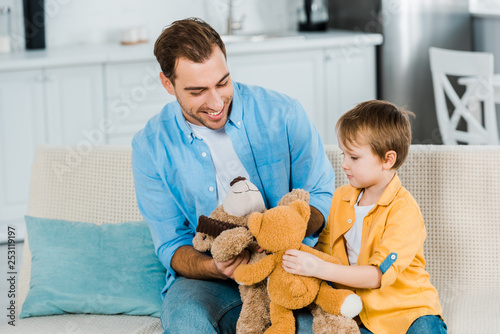 father and preschooler son sitting on couch and playing with teddy bears at home