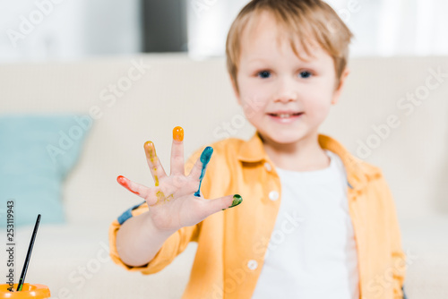 cute preschooler boy with colorful paint on hand looking at camera at home