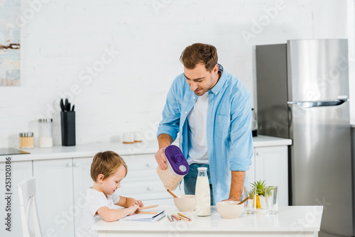 handsome father pouring cereal in bowl while preschooler son drawing during breakfast in morning