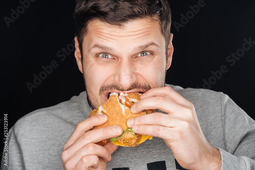handsome man eating tasty chicken burger while looking at camera isolated on black