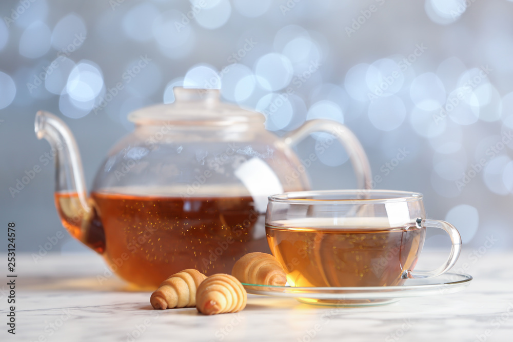 Glass cup and teapot with black tea on table against blurred lights