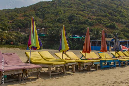 multicolored old beach beds with umbrellas on the sand against a green hill with huts