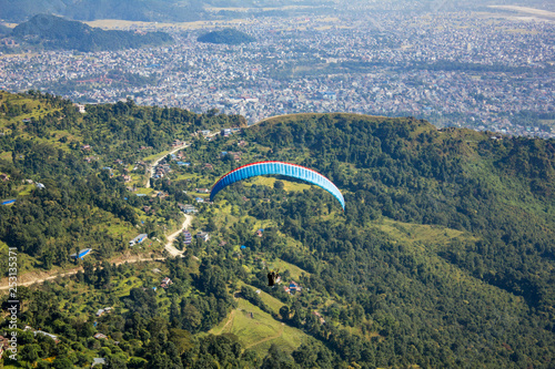 paraglider flies on a blue parachute against a green mountain and a city in the valley