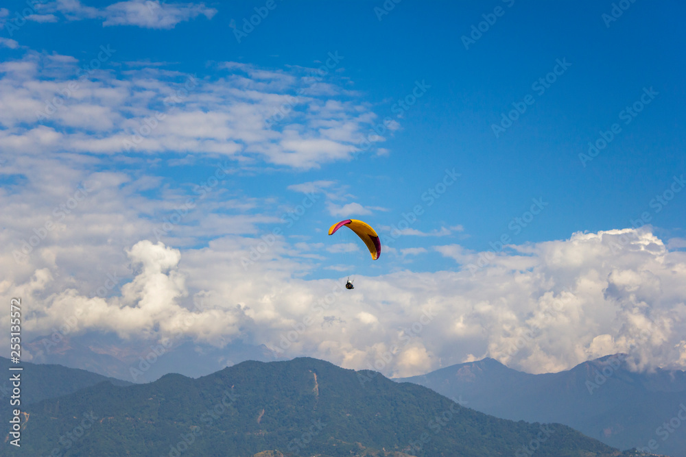 paraglider flies on a yellow parachute against a green mountain and blue sky with white clouds