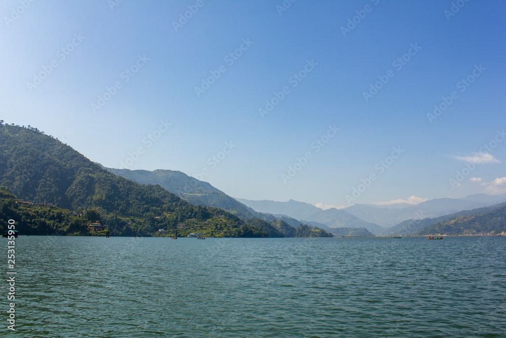 Phewa lake with boats on the background of a green mountain valley under the blue sky, view from the water