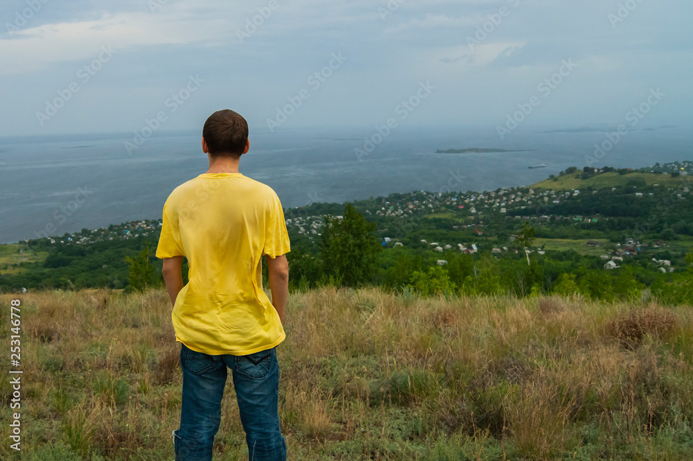 The guy stands on a mountain and looks into the distance to the village