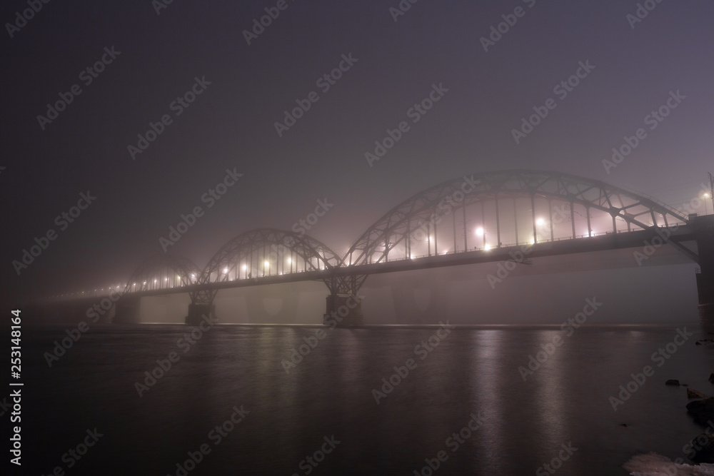 A mysterious evening fog above the river in big city. Bridge in the mist, cold weather scenery. Soft, blurry, misty look. Colorful, mystic industrial cityscape. A mysterious evening fog above the river in big city. Bridge in the mist, cold weather scenery. Soft, blurry, misty look. Colorful, mystic industrial cityscape.