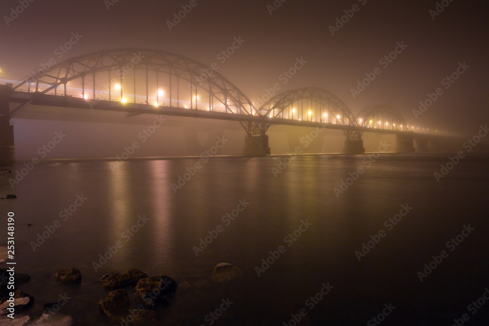 Fototapeta premium A mysterious evening fog above the river in big city. Bridge in the mist, cold weather scenery. Soft, blurry, misty look. Colorful, mystic industrial cityscape.