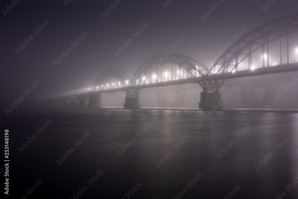Obraz premium A mysterious evening fog above the river in big city. Bridge in the mist, cold weather scenery. Soft, blurry, misty look. Colorful, mystic industrial cityscape.