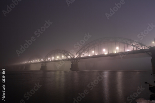 A mysterious evening fog above the river in big city. Bridge in the mist, cold weather scenery. Soft, blurry, misty look. Colorful, mystic industrial cityscape.