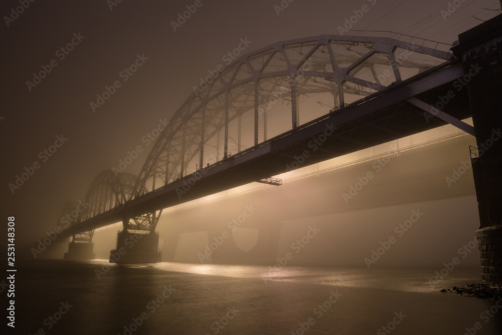 Obraz premium A mysterious evening fog above the river in big city. Bridge in the mist, cold weather scenery. Soft, blurry, misty look. Colorful, mystic industrial cityscape.