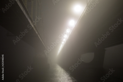 A mysterious evening fog above the river in big city. Bridge in the mist, cold weather scenery. Soft, blurry, misty look. Colorful, mystic industrial cityscape.