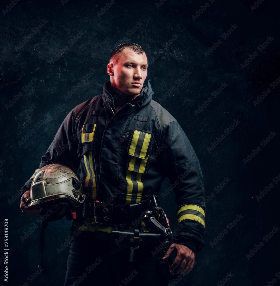 Brutal firefighter looks sideways and holds a helmet in his hand in the studio on a black background Brutal firefighter looks sideways and holds a helmet in his hand in the studio on a black background