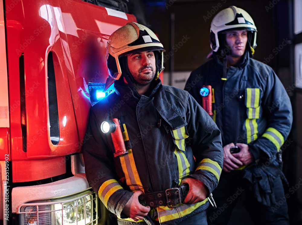 Two firemen in the fire station garage, standing next to a fire truck and looking sideways Two firemen in the fire station garage, standing next to a fire truck and looking sideways